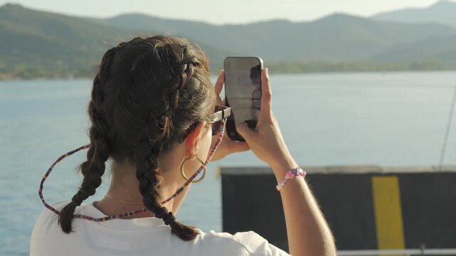 Young woman photographing Elba island sunset from ferry, Italy