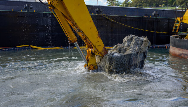 Excavator digging in water on sea shore or bay, Excavator bucket scoops stone boulders sand near pier dock