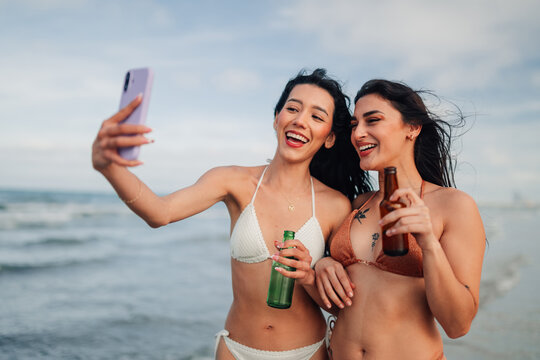 Happy young women friends taking beach selfie on summer vacation, laughing and enjoying beer outdoors