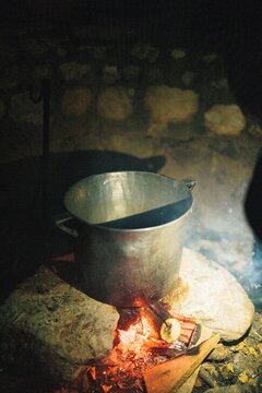 A metal pot sits over glowing embers, steam rising into the cool night air. Shadows dance on the stone walls of a cozy cabin, creating a warm, inviting atmosphere perfect for storytelling
