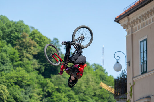 Mountain biker performing inverted backflip trick during freestyle competition in urban setting, extreme cycling action showcasing skill, risk and athletic performance