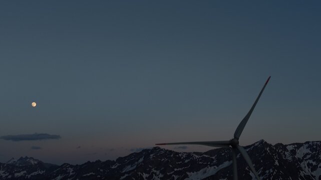 Aerial view of a wind turbine at the Gotthard Wind Farm with a full moon over snow-capped mountain peaks during twilight in Airolo, Ticino, Switzerland.