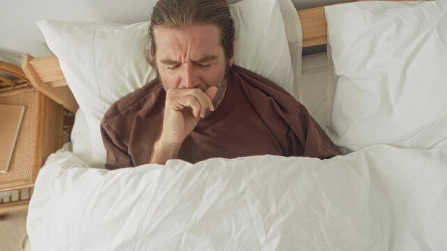 Man covering mouth yawning in bed under white duvet with pillow on bedside table inside a building; fatigue morning rest contemplation.