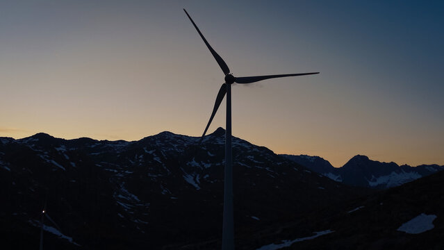 Aerial view of the Gotthard Wind Farm silhouette against a sunset sky and snow-capped mountains in Airolo, Ticino, Switzerland.