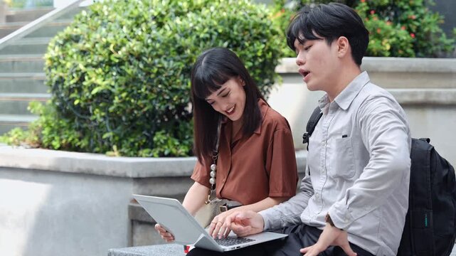 Asian business professional student sitting outdoor on stair holding laptop, woman smiling and talking with male classmate with backpack during college project research