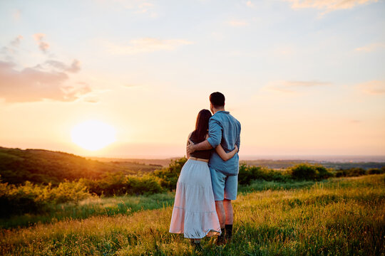 Young couple standing in a field at golden hour, romantically embracing and watching the sunset over rolling hills