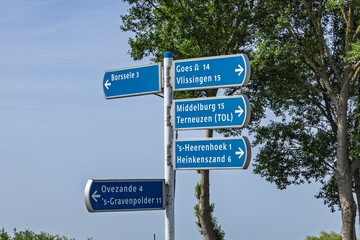 Fototapeta premium Zeeland, Netherlands. Blue directional signpost indicating distances to local towns like Borssele, Goes, Middelburg, Vlissingen, and Terneuzen under a clear blue sky.