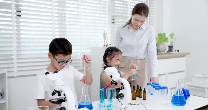 Asian boy and girl age around 6 smiling and working with test tubes filled with blue liquid beside educator during elementary science class experiment using microscope and lab equipment