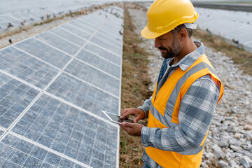 Obraz premium Worker checks panels with tablet, wearing hard hat and vest. Focused, calm, professional attitude evident. Solar farm under clear sky, rows of panels stretch wide. Ideal for renewable energy
