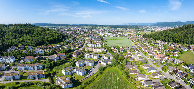 Aerial view of residential neighborhoods, apartment buildings with solar panels, and green fields surrounded by forested hills in Zofingen, Aargau, Switzerland.