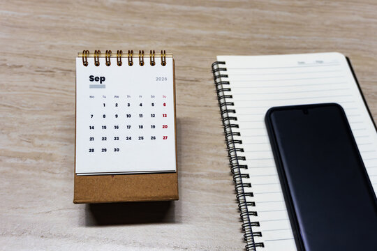 Top view of September 2026 desk calendar with smartphone and blank spiral notebook on wooden table. Copy space for time management, event planning, or schedule concept.