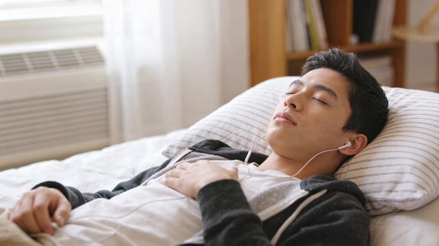 Audio starting lulling student in zip-up hoodie lying on bed in bedroom, breathing with earphones