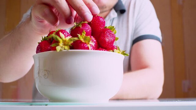 Low angle close-up of man's hands eating ripe strawberries from a bowl, blurred silhouette background. Shallow depth of field, natural window light.Healthy nutrition -organic food. 