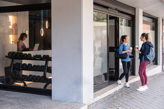 Two women chatting outside gym while trainer works inside