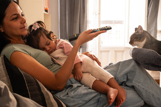 Latin mother using remote while holding baby daughter near cat at home
