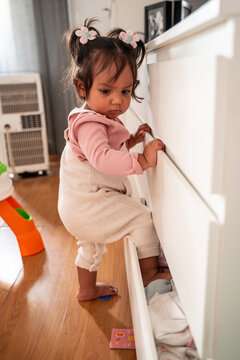 Latin baby girl climbing open dresser drawer at home