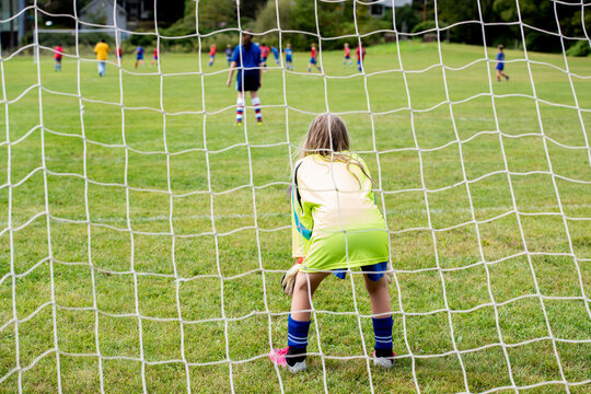 Young girl plays goalkeeper during a youth soccer match, crouching low behind the white goal net as she watches the action unfold at the far end of the field. 
