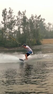 Vertical video: Wakeboarding man being towed by cable at lake holding cable doing trick with helmet