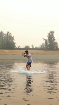 Vertical video: Cable towing rider from water onto wakeboard, carving past box on lake in helmet