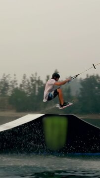 Vertical video: Carving rider in helmet, cable pulling to kicker on lake, launching, splashing