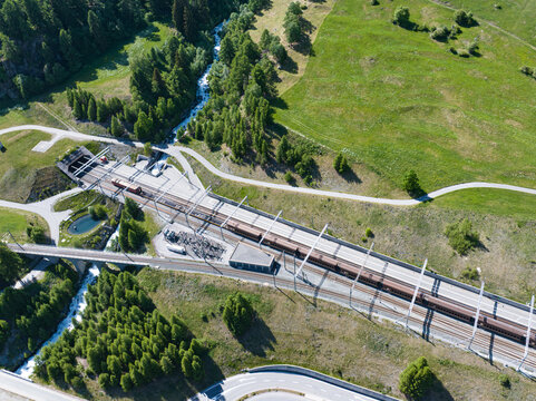 Aerial view of the Vereinatunnel Klosters railway entrance with a train on the tracks, surrounded by lush green meadows, pine forests and a mountain stream Zernez, Grisons, Switzerland.