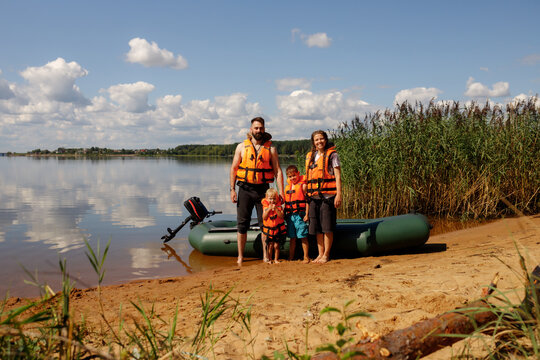 Happy family with two kids wearing orange life jackets standing by green inflatable boat on sandy beach of river or lake, active summer weekend, outdoor water recreation, security safety concept