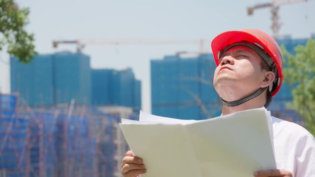 During sweltering summer heat, a sweaty construction engineer with a safety helmet checks construction blueprints on the building site.