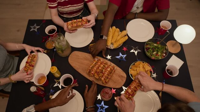 Overhead view of multiethnic young adult friends reaching for hot dogs on decorated table during Independence Day celebration. Clip is perfect for holiday marketing and food advertisements