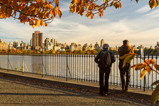 Senior travelers standing on a park path looking at the city skyline and reservoir during autumn, reflecting on a tourism adventure in Central Park, Manhattan, New York City, USA.
