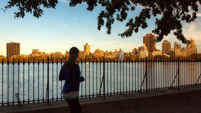 Female runner exercising along a reservoir path, representing an active urban lifestyle during golden hour in Central Park, Manhattan, New York City, United States.