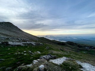 A wide landscape view of rugged rocky pastures on a mountain slope, looking towards a soft golden sunrise over distant peaks blanketed in low clouds. © Licia.Galli