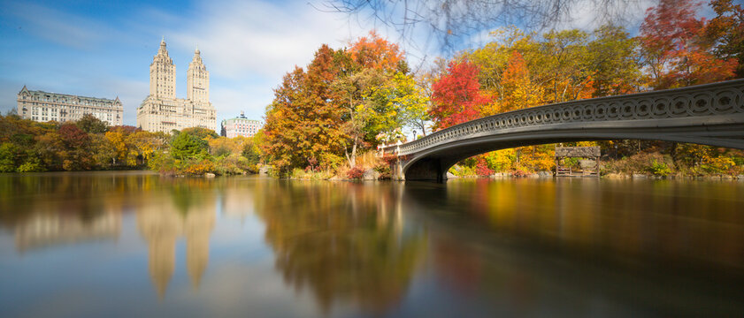 Bow Bridge reflects over The Lake amidst colorful autumn foliage and urban skyline, highlighting an iconic travel and tourism destination in Central Park, Manhattan, New York City, USA.