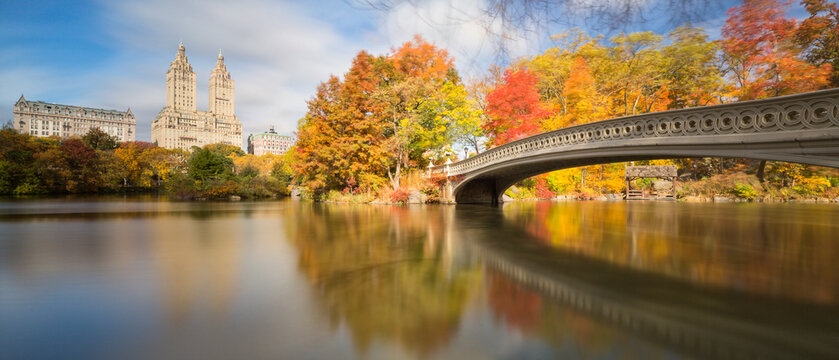 Bow Bridge spans the calm lake reflecting vibrant autumn foliage and urban skyscrapers, symbolizing tourism and travel in Central Park, Manhattan, New York City, United States.