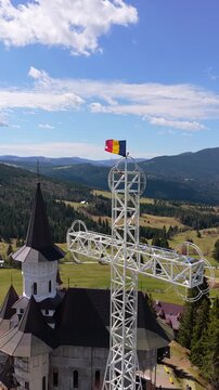 Aerial drone view of a Romanian flag waving above the large metal cross near Acoperamantul Maicii Domnului Monastery with the mountains in the background. Vertical