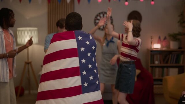 Medium portrait of African American young adult man dancing with USA flag during indoor party with multiethnic friends. Footage is ideal for Independence Day promotions and patriotic holiday campaigns