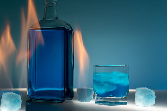 Bright orange flames rise behind a glass bottle next to a tumbler glass filled with blue liquor and ice cubes on a white surface against a blue gradient background