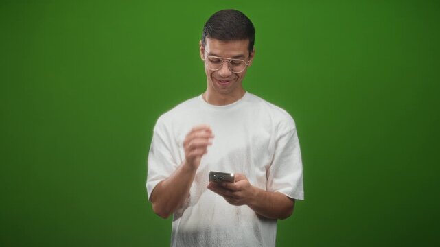 Man holding smartphone with finger to chin in studio, smiling while reading a message on his phone, wearing white tshirt and clear glasses; playful curiosity.