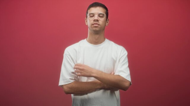 Man adjusting sleeve and touching upper arm in studio with red wall, wearing glasses and a white tshirt; quiet contemplation.