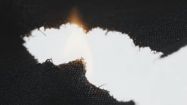 Burning black woven textile growing flame from small orange glow at studio macro, with backlight