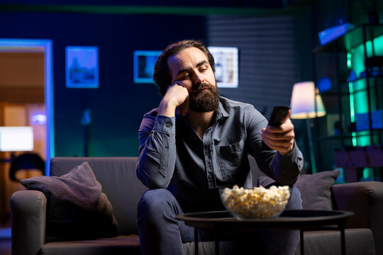 Cord cutter bored at home using TV to watch films on streaming services, zapping through content. Man in living room mindlessly consuming entertainment, using remote control