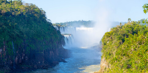 A breathtaking panoramic view of the majestic Iguazu Falls. Massive cascades rush through the canyon, creating a dramatic mist. The surrounding lush, vibrant green jungle cliffs highlight the scale. © Ilya