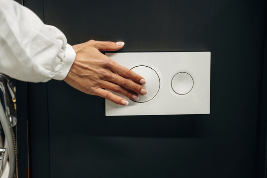 Hand of a person with manicured nails pressing a modern dual flush toilet button on a sleek white panel against a dark wall in a contemporary bathroom setting
