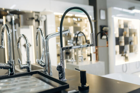 Modern kitchen faucet displayed on a countertop with various other faucet designs and fixtures visible in a well-lit showroom environment
