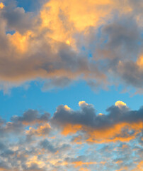 Obraz premium Premium vertical view of a beautiful dramatic sky filled with thick sunset clouds illuminated by bright orange golden hour light against a calm blue background.