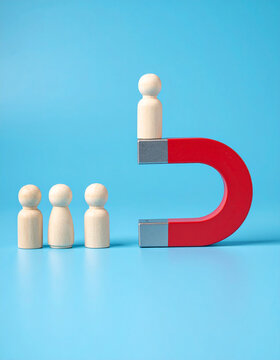 Large red magnet attracting wooden figure from row on blue background, illustrating talent acquisition and lead generation, modern corporate concept. Close-up.