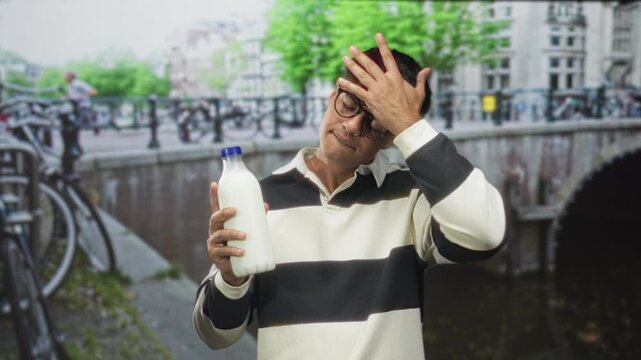 Man holding milk bottle with hand on forehead gesture on street bridge by canal, wearing glasses and striped shirt; forgetfulness frustration.