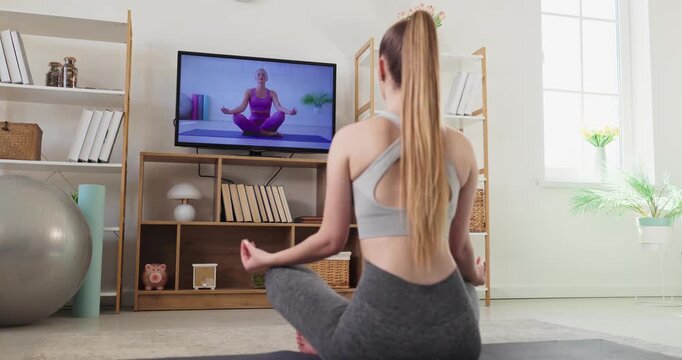 Woman yoga home meditation online fitness workout. She follows an online class on a television, mirroring a pose. Calm focus and steady breathing show balance. Clear mindful wellness at home