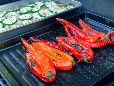 Fresh red pointed peppers and seasoned zucchini slices cooking on a barbecue grill. The scene captures healthy, vegan outdoor cooking during a bright summer day.