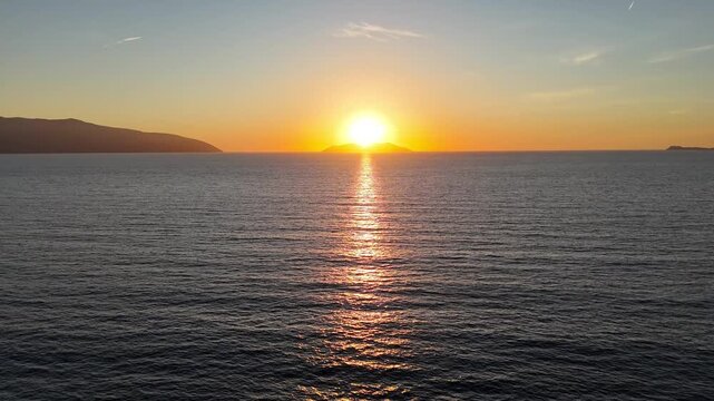 Aerial view of sunrise over calm sea waves, with golden sunlight reflecting on water surface and distant mountains silhouetted against the horizon in a serene coastal landscape. Sunset over the ocean