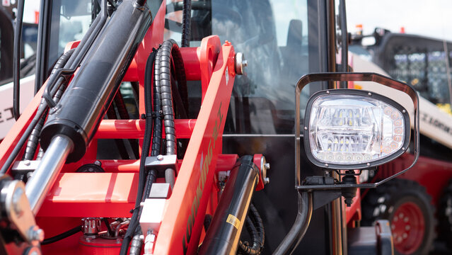 Detailed close-up of red farm machinery showing hydraulic cylinders, metal arms, hoses, and a front work light beside the cab, with industrial equipment visible in the background